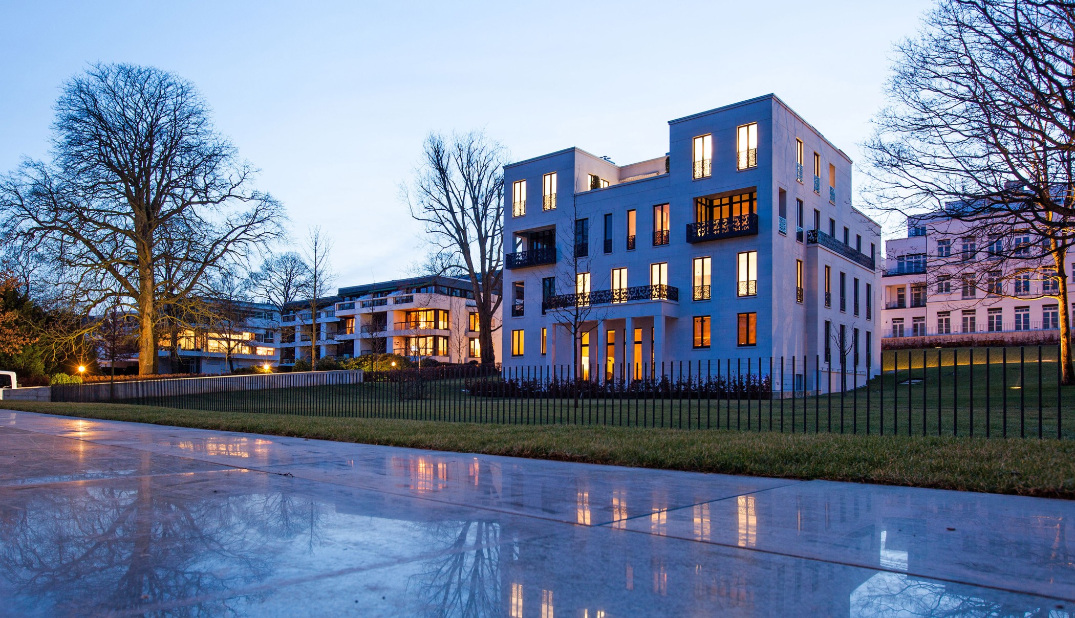 Alster villas at dusk with lit windows, bare trees and a reflective paved square