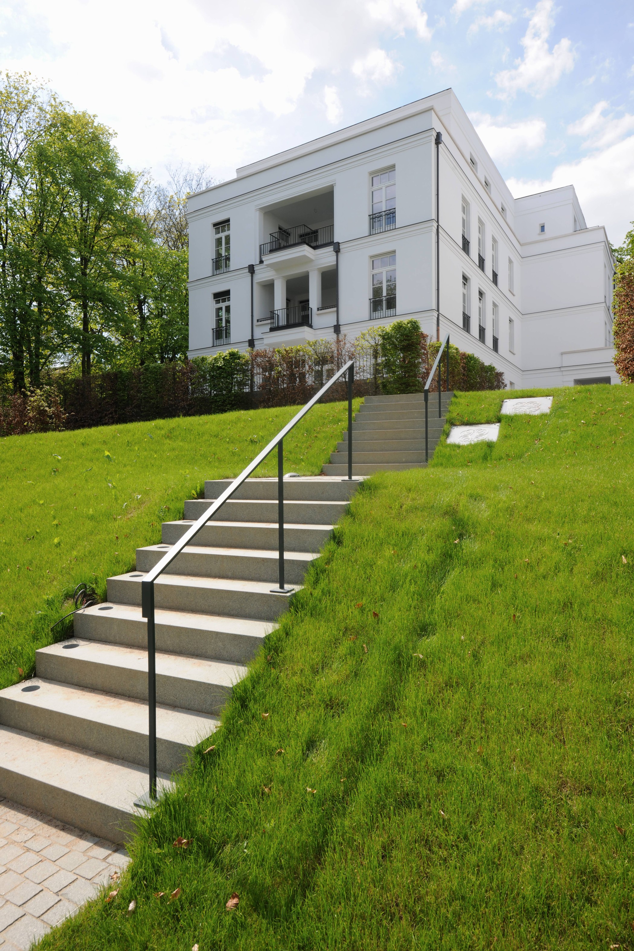 Treppe mit Metallhandlauf an einem Rasenhang, die zu einer weißen Villa der Alstervillen hinaufführt