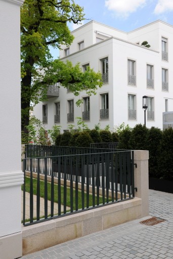 Weiße Wohnhäuser mit Laternen hinter einem dunklen Metallzaun und Hecke, davor ein gepflasterter Gehweg