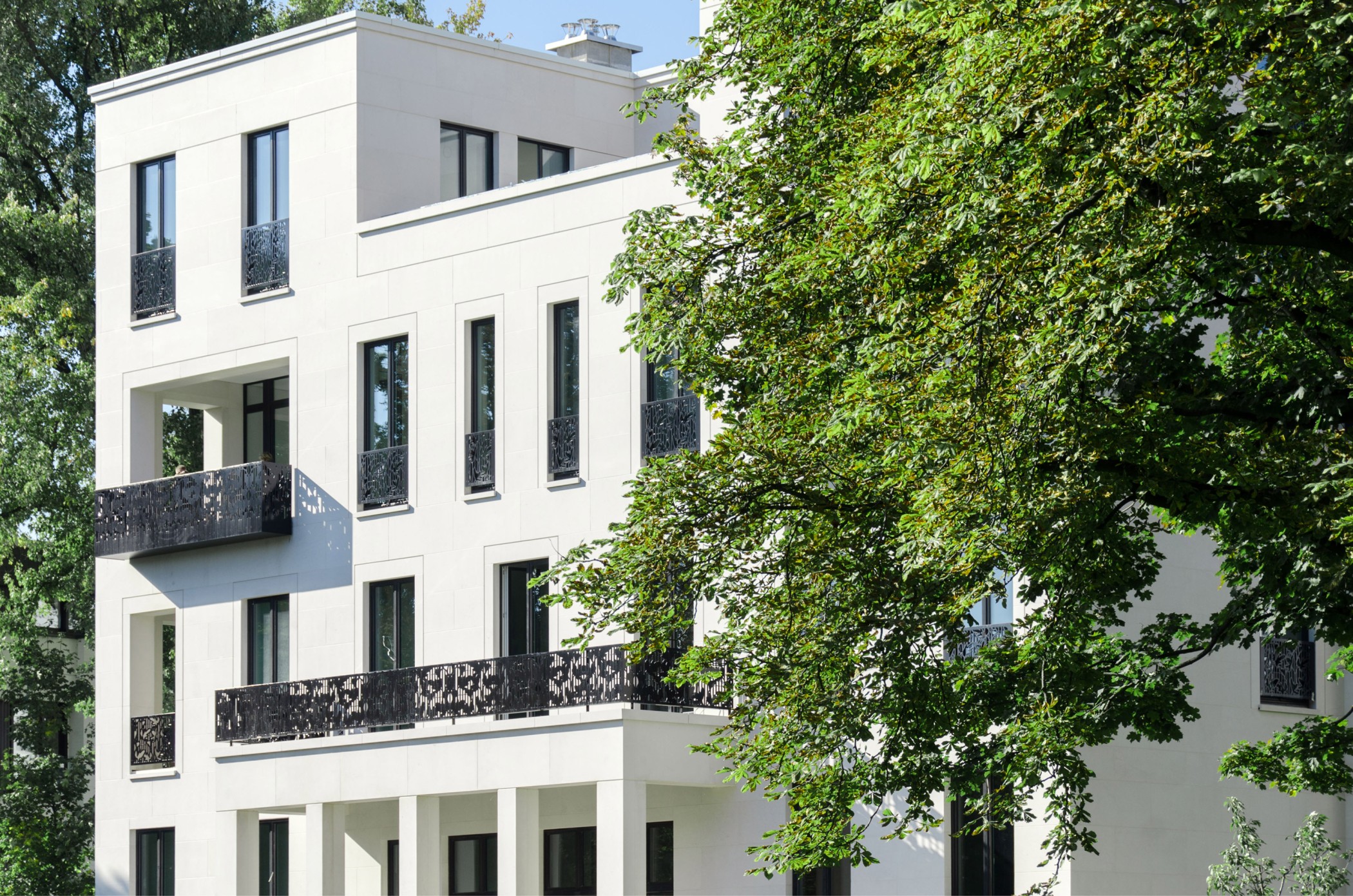 Weiße Fassade der Alstervillen mit schwarzen ornamentalen Balkongeländern, daneben ein großer Laubbaum